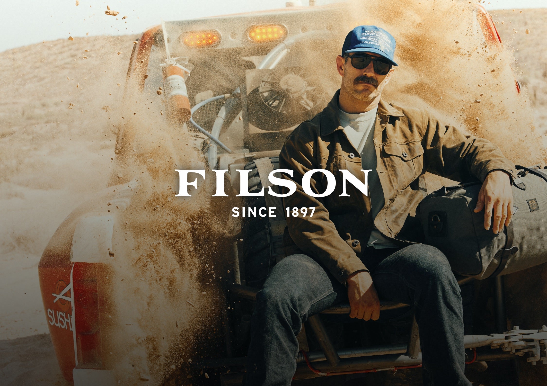 A man wearing a blue FILSON cap, sunglasses, and a brown jacket sits on the front of a dusty off-road vehicle in a desert setting. The FILSON brand logo is prominently displayed in the center of the image.