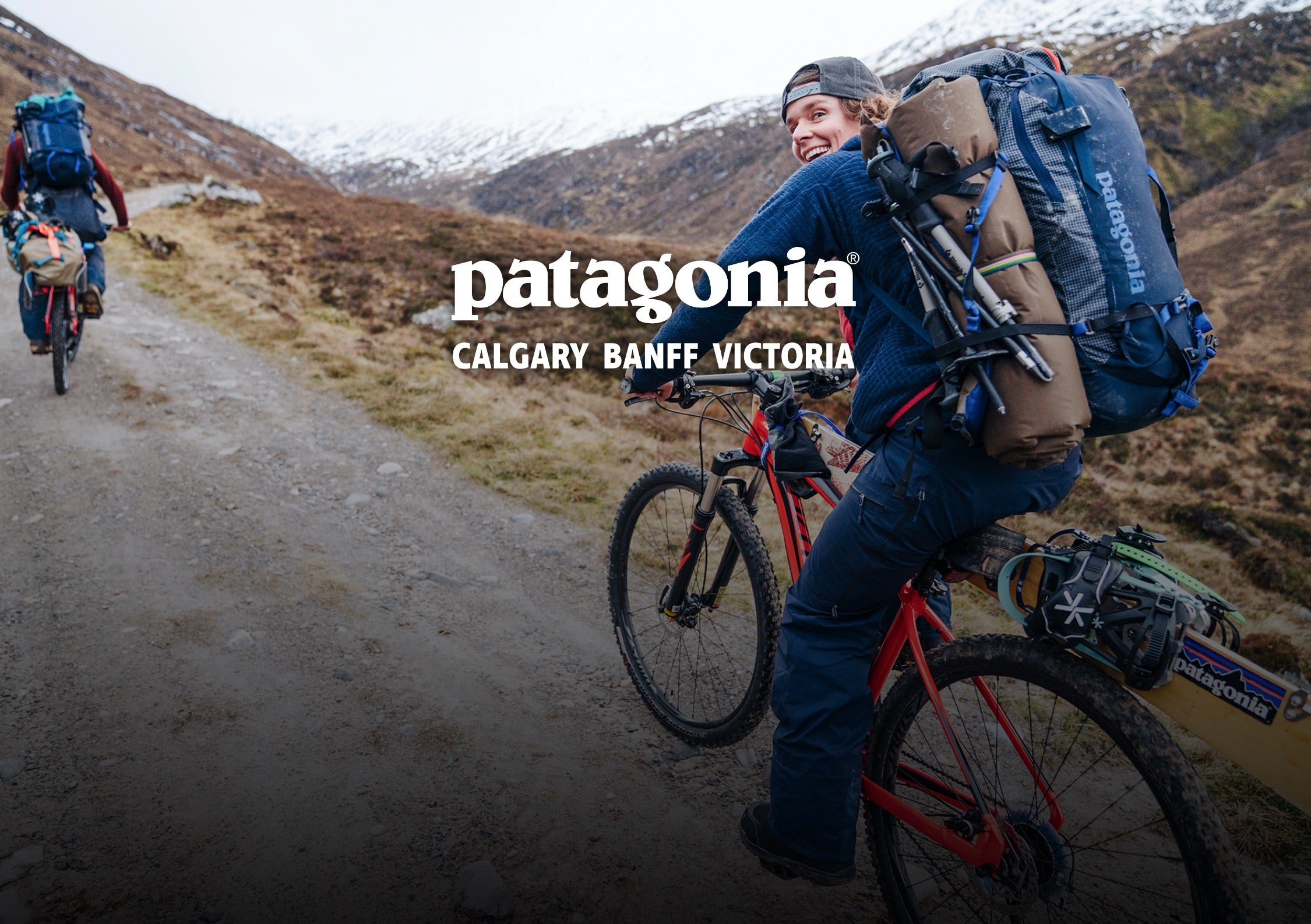 A man on a red mountain bike rides along a dirt trail in a mountainous outdoor setting, wearing a blue Patagonia backpack and helmet. Another cyclist is visible in the background.